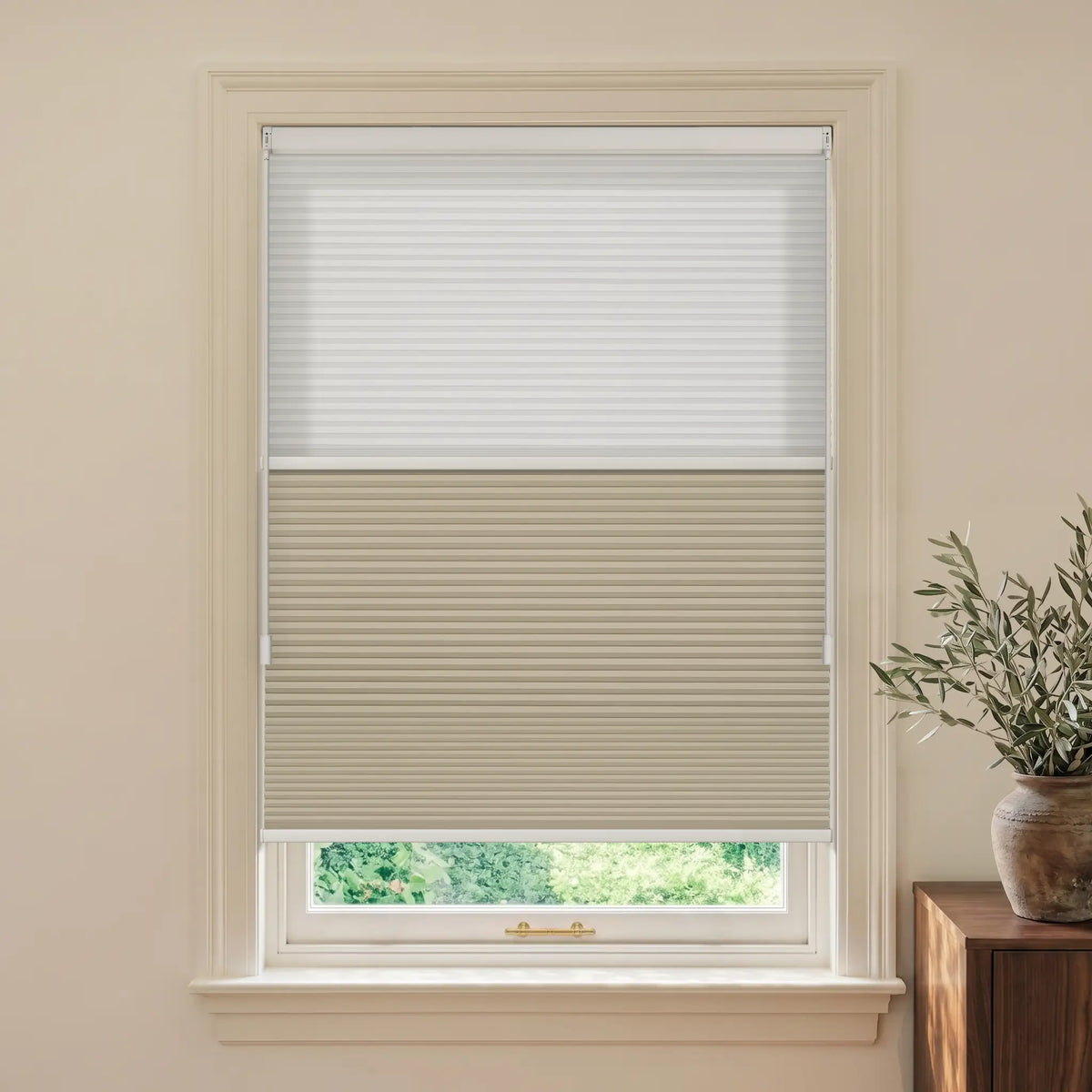 A window with beige and white Motionblinds smart Day Night Cellular Shades partially drawn, set in a cream-colored wall with a wooden side table and potted plant visible on the right.