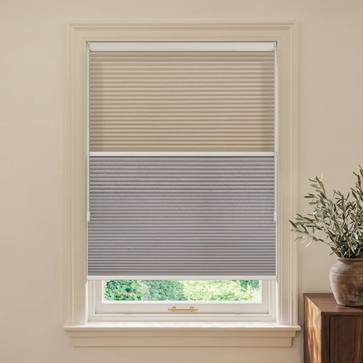 A Motionblinds smart Day Night Cellular Shades  in beige and gray shown installed on a white-framed window in a neutral beige room, with a small potted plant on a wooden side table to the right.
