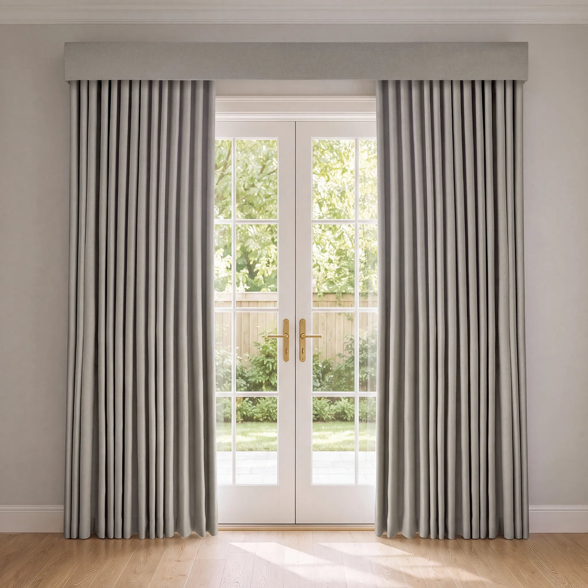 A pair of light gray pleated window curtains hanging on either side of white French doors, with a matching gray valance at the top. The room has light-colored walls and a light wood floor, with natural light coming from the doors showing green garden foliage outside.