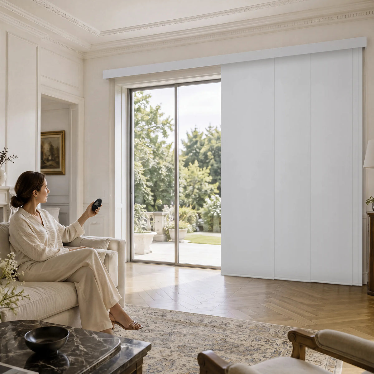 A woman in a cream lounge set sits on a sofa in a bright, elegant living room, holding a remote control near a large sliding glass door. A white Motorized Blackout Panel Track Blinds is partially open, revealing a garden outside.