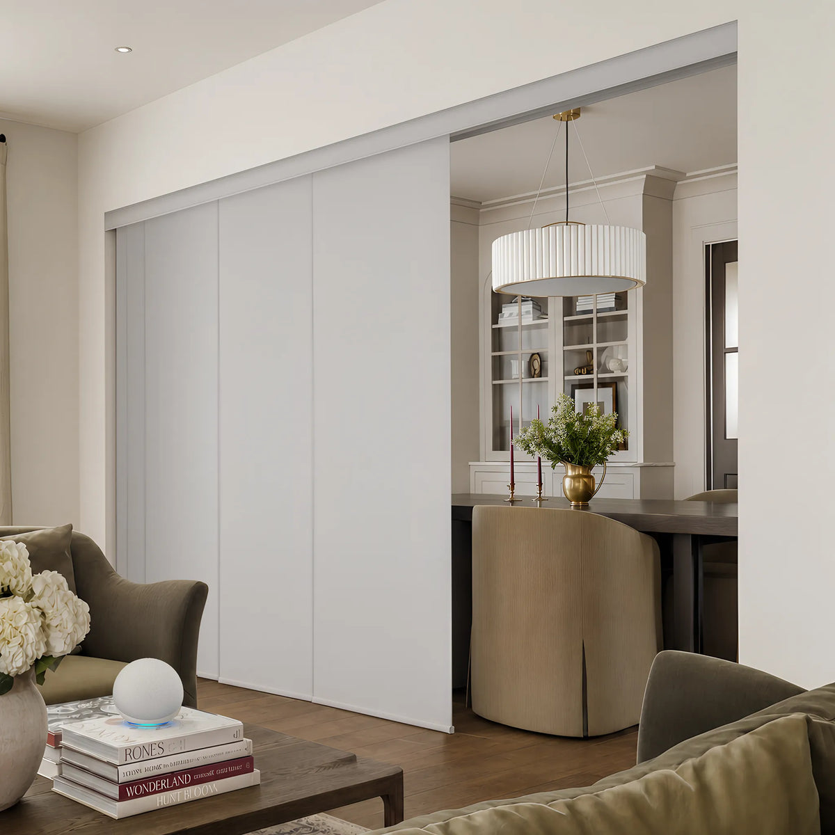 A modern interior scene showing a Motorized Blackout Panel Track Blinds installed in a doorway, with a dining area and shelving visible behind it. The room includes upholstered seating, a coffee table with books and flowers, and a pendant light.