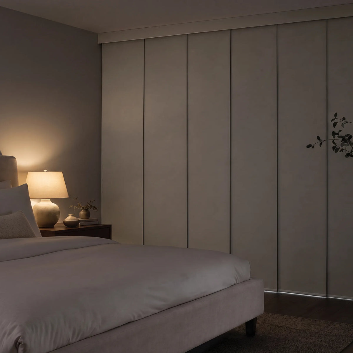 A modern bedroom scene featuring a Motorized Blackout Panel Track Blinds  in a warm taupe-beige finish, styled with a neatly made bed, a lit bedside lamp, and a dark wood nightstand.