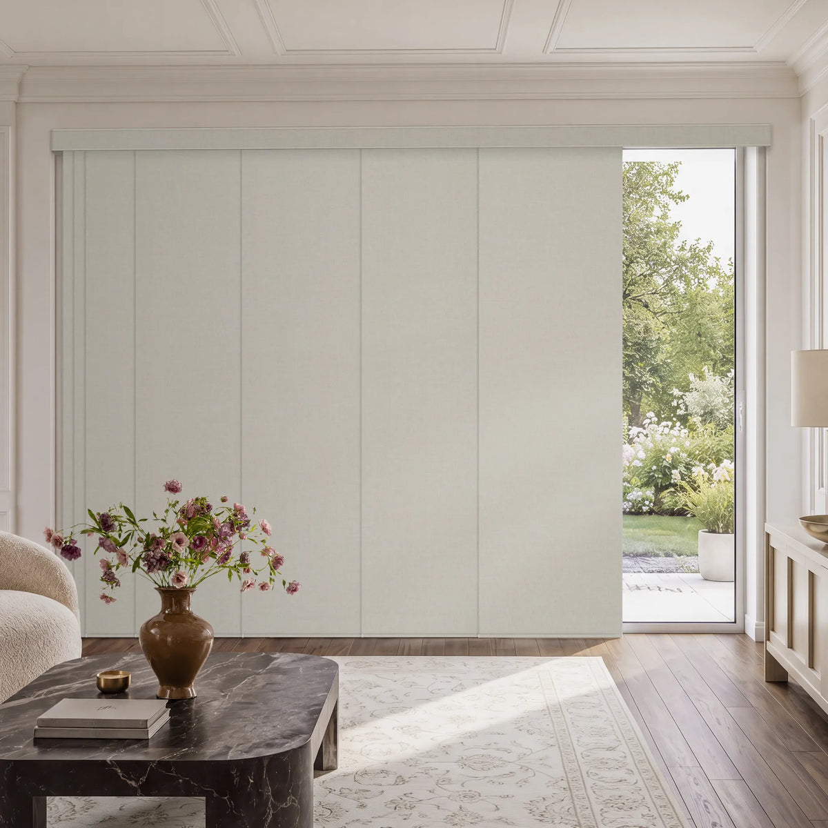 A large sliding panel door in a warm off-white or cream color installed in a white paneled room, with a dark marble coffee table, a vase of flowers, and a view of a garden visible through an open panel on the right.