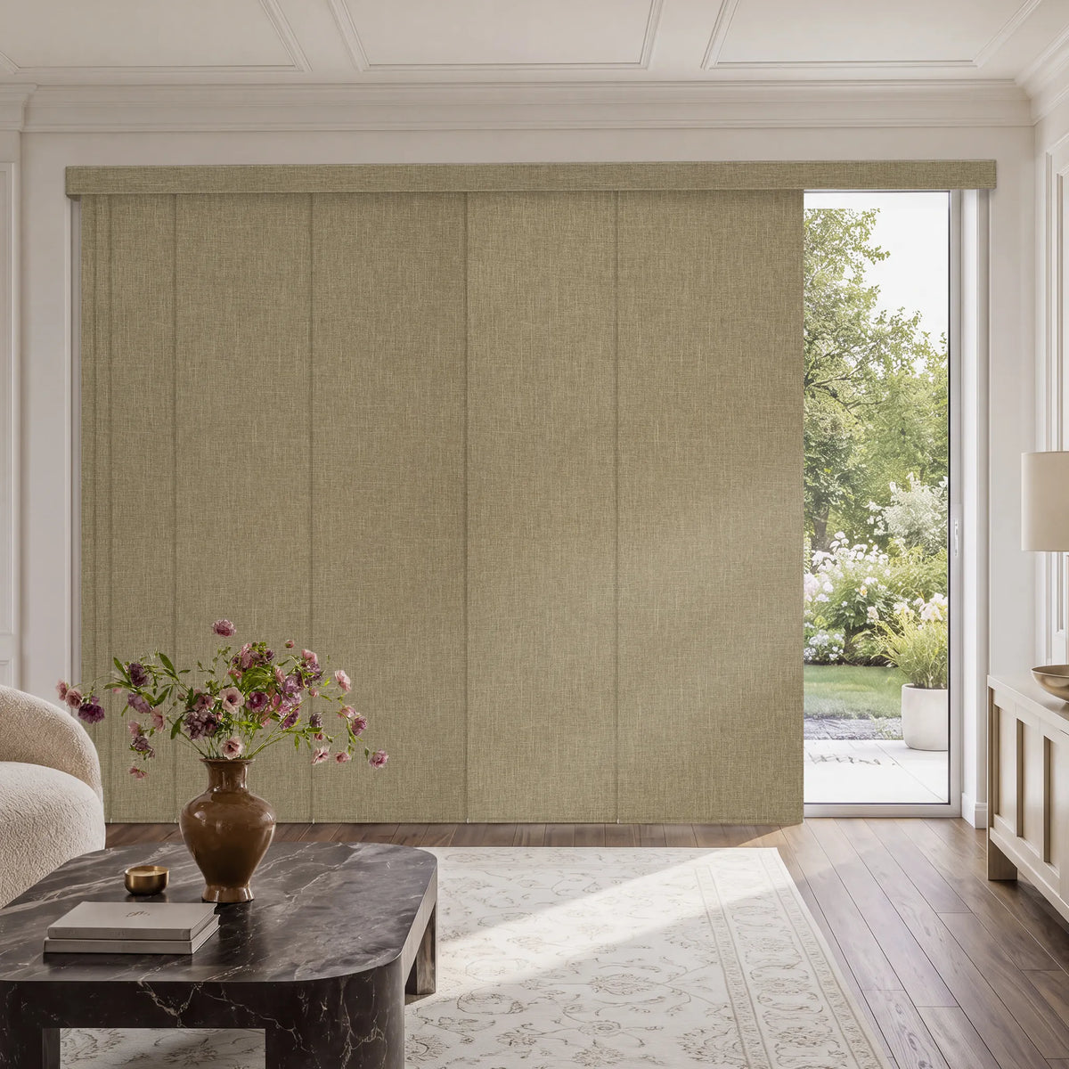 A large beige Motorized Blackout Panel Track Blinds  covers most of a sliding glass door in a bright, elegant living room with a marble coffee table, vase of flowers, and outdoor garden view.