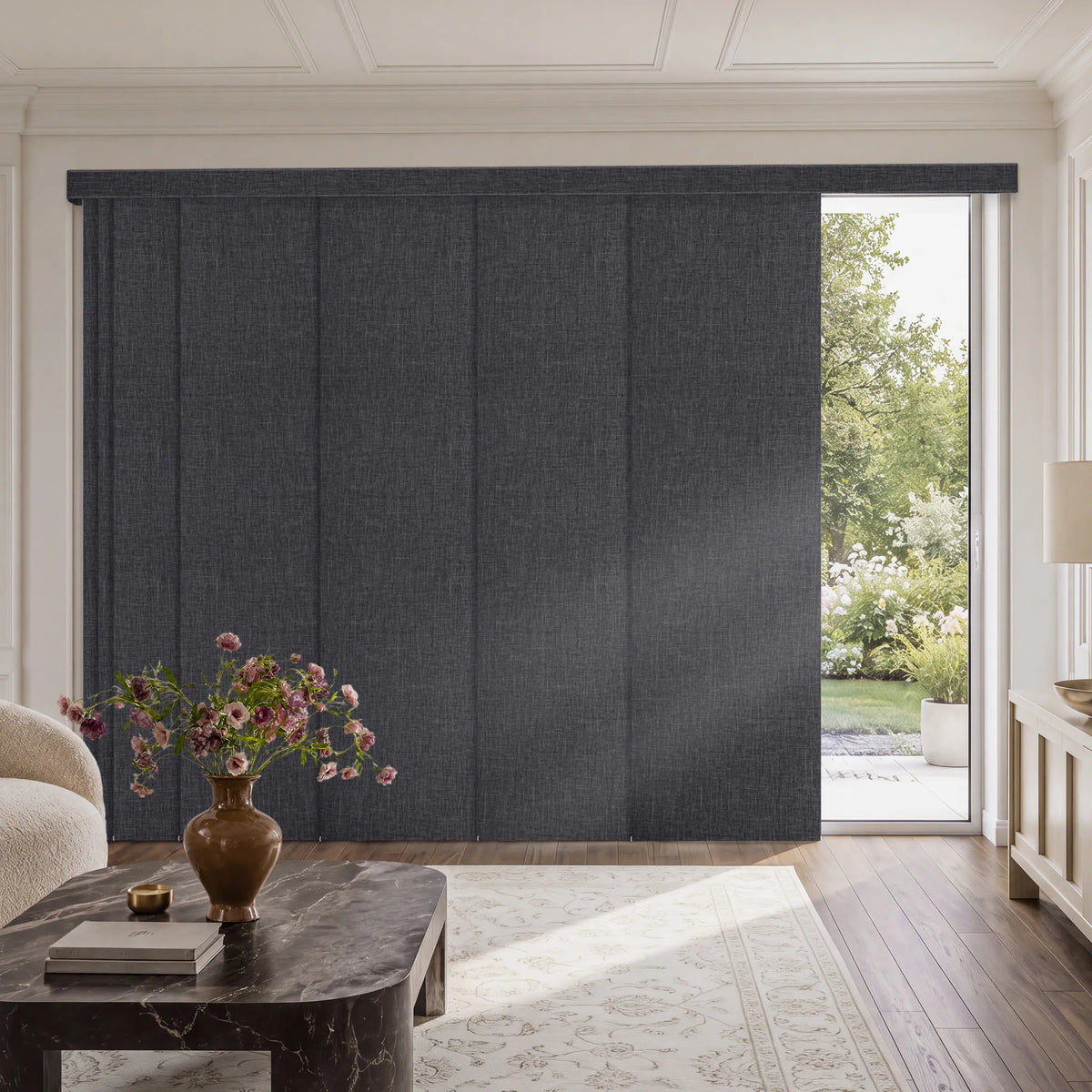 A large dark charcoal gray textured Motorized Blackout Panel Track Blinds  installed in a bright, elegant living room with hardwood floors, a marble coffee table, and a garden view visible through an open section.