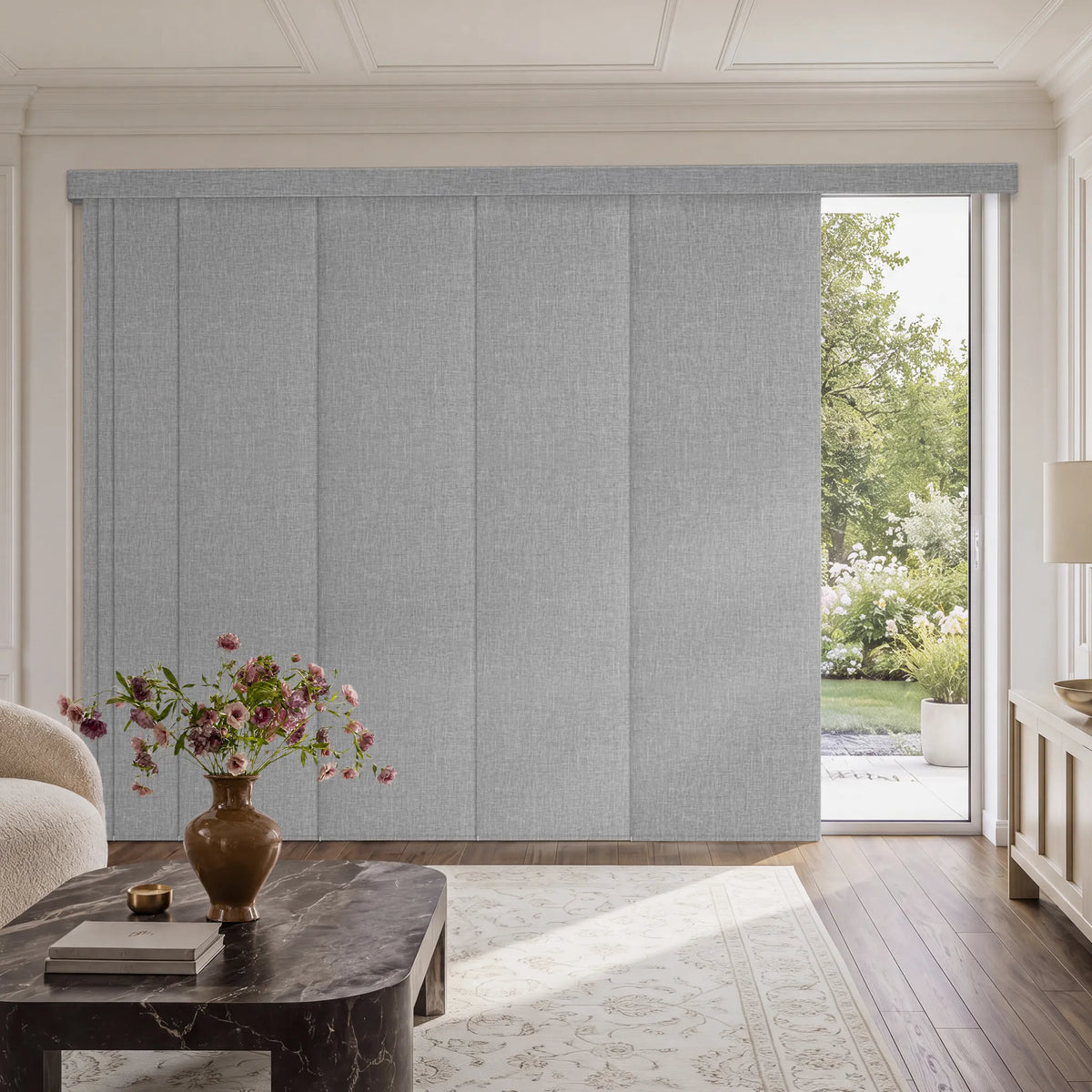 Light gray Motorized Blackout Panel Track Blinds  installed in a bright, modern living room with a marble coffee table, floral arrangement, and a garden view visible through an open window.