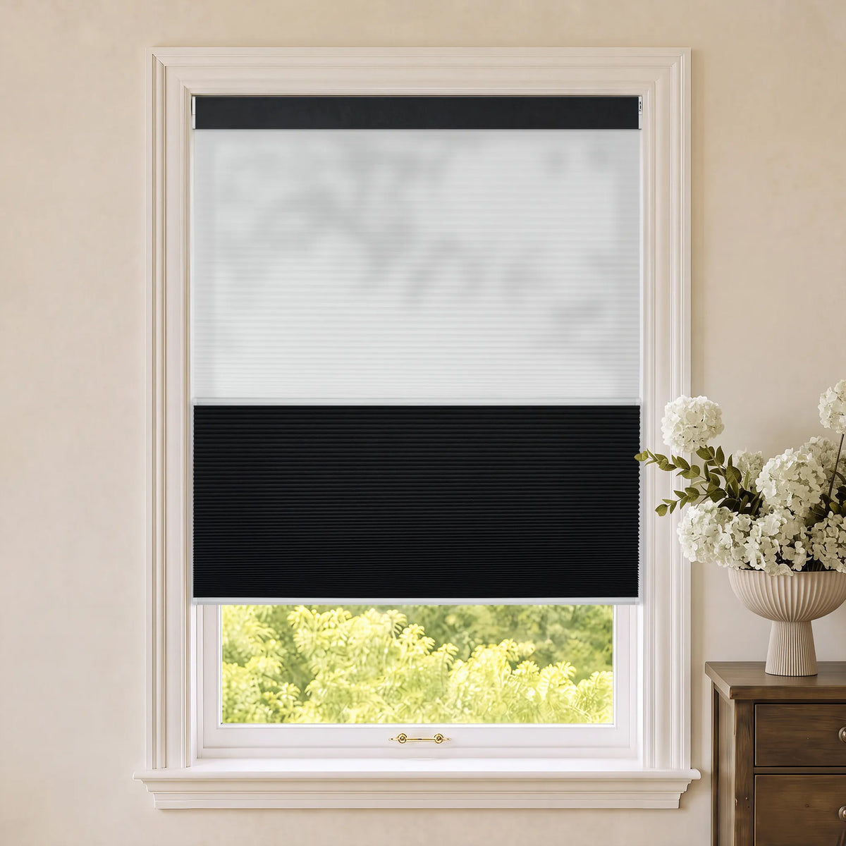Motorized Day Night Cellular Shades， shown installed on a window in a beige room with a vase of white flowers on a wooden side table.