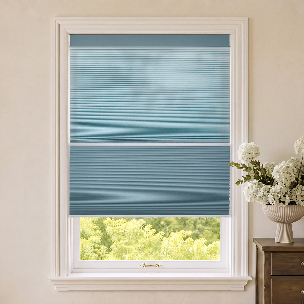 A two-tier blue Motorized Day Night Cellular Shades installed on a white-framed window, with a view of green trees outside and a vase of white flowers on a wooden side table to the right.