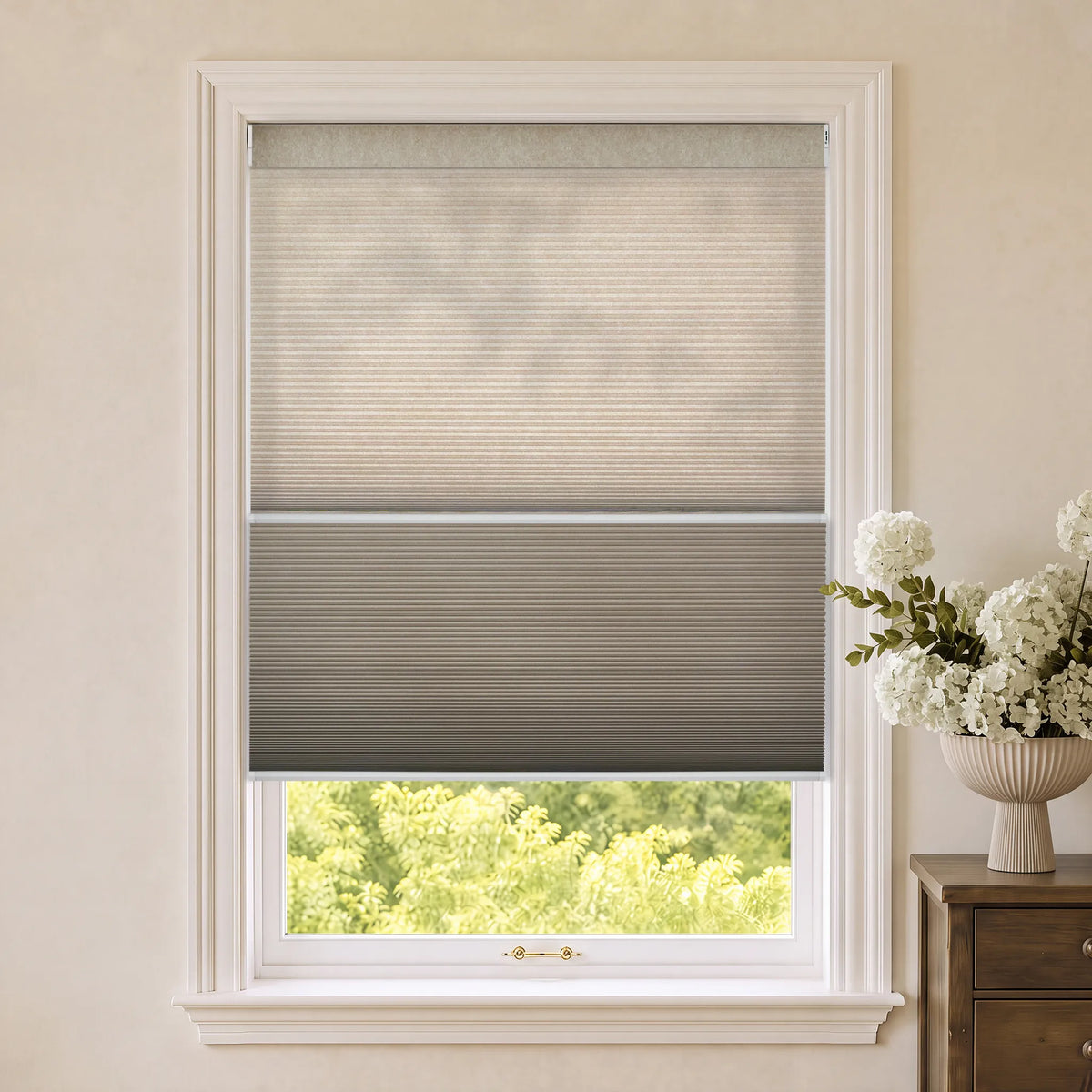 Motorized Day Night Cellular Shades partially rolled down on a white-framed window, with a vase of white flowers on a wooden side table next to it.