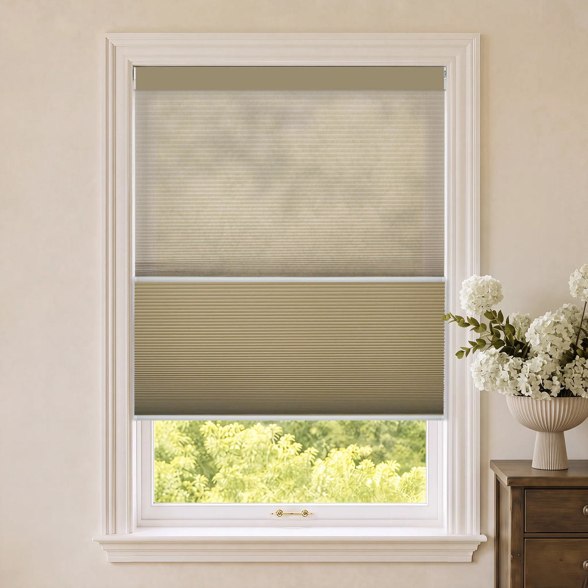 A Motorized Day Night Cellular Shades partially drawn on a white window, set in a warm beige room with a wooden side table and white flowers visible on the right.