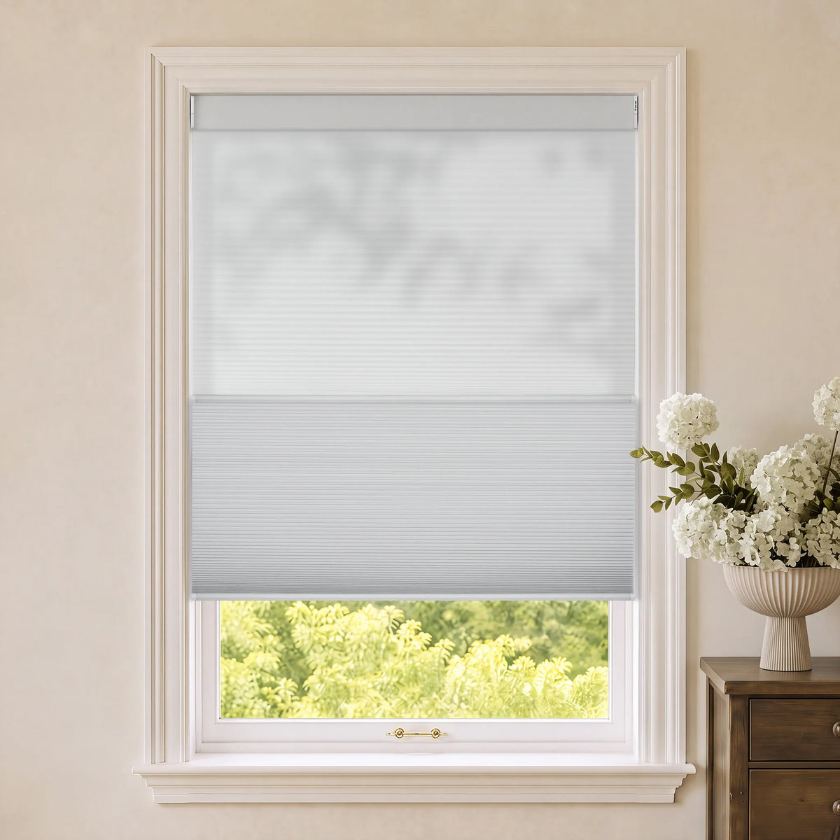 A light gray Motorized Day Night Cellular Shades partially rolled down in a white-framed window, set in a beige room with a vase of white flowers on a wooden side table.