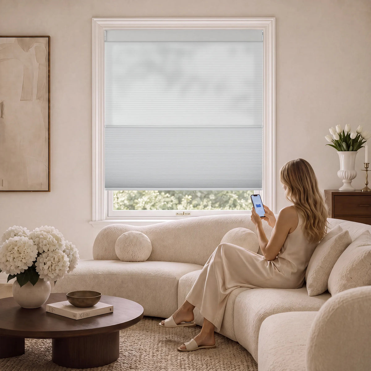 A woman sits on a cream sectional sofa in a bright, modern living room, looking at her phone. The product being showcased is the light gray Motorized Day Night Cellular Shades installed on the window behind her.