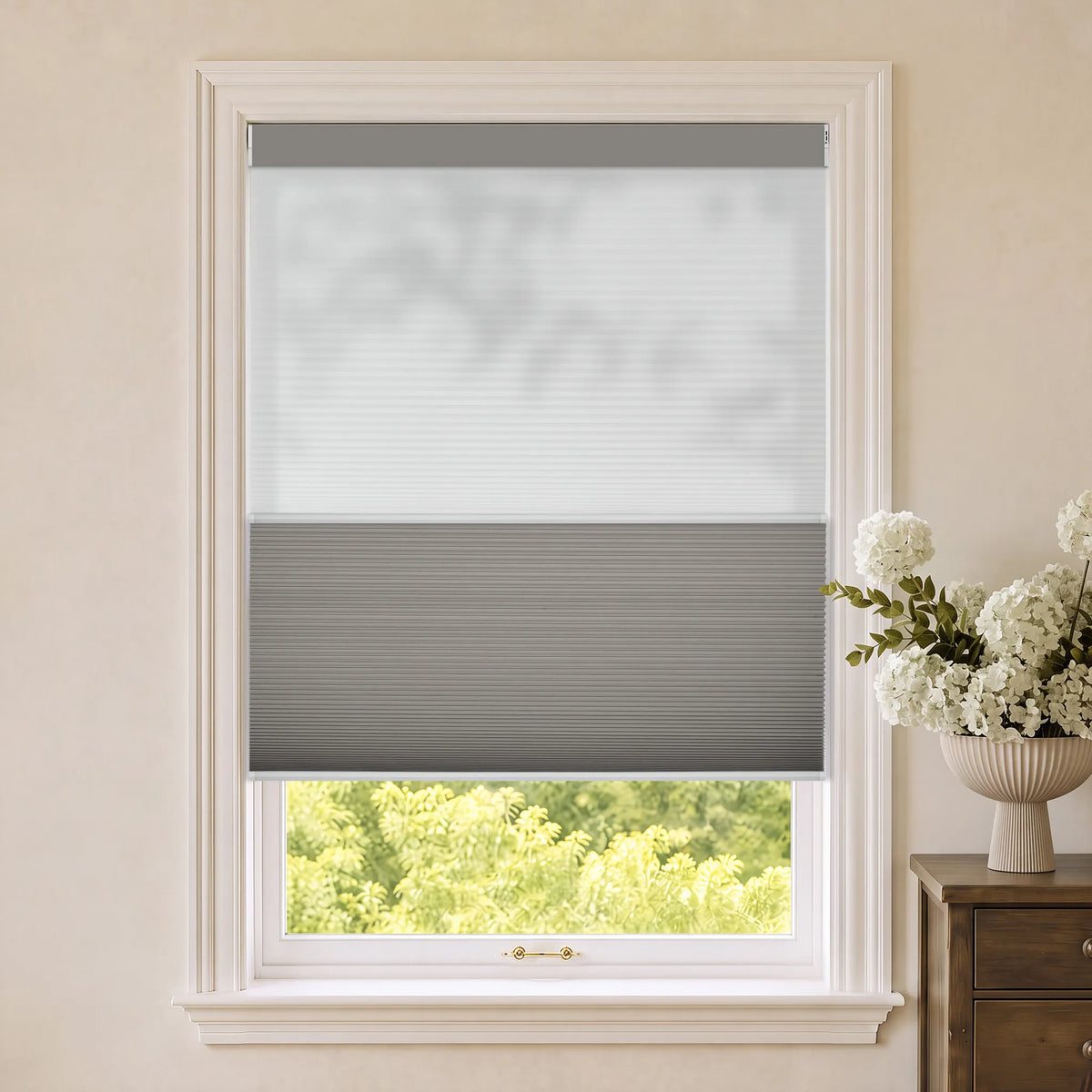 Gray Motorized Day Night Cellular Shades installed in a white-framed window, shown with a beige wall and green foliage visible outside, alongside a wooden side table with white flowers.