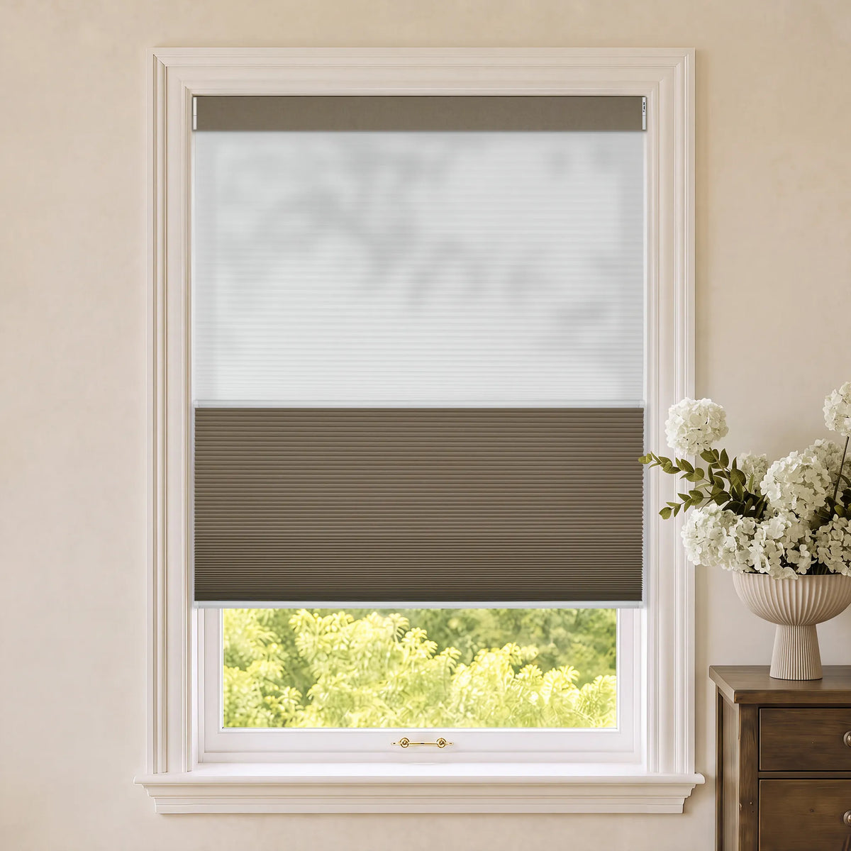 Motorized Day Night Cellular Shades, installed on a white-framed window in a beige room with a vase of white flowers on a wooden side table.