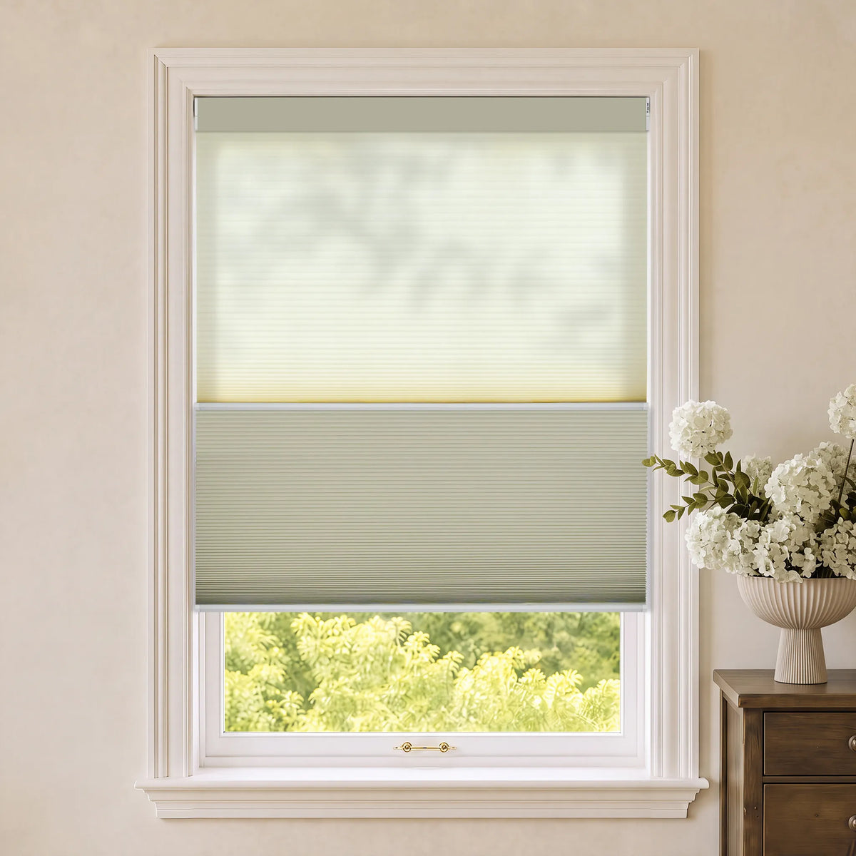 A Motorized Day Night Cellular Shades with a light sage green top section and a beige bottom section, shown installed on a white-framed window in a neutral-toned room with a vase of white flowers on a wooden side table.