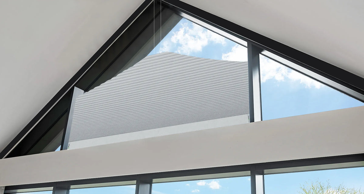 A close-up view of a white Motorized Elongated Triangle Cellular Shades partially drawn, installed inside a modern building with a large triangular skylight and black frames.