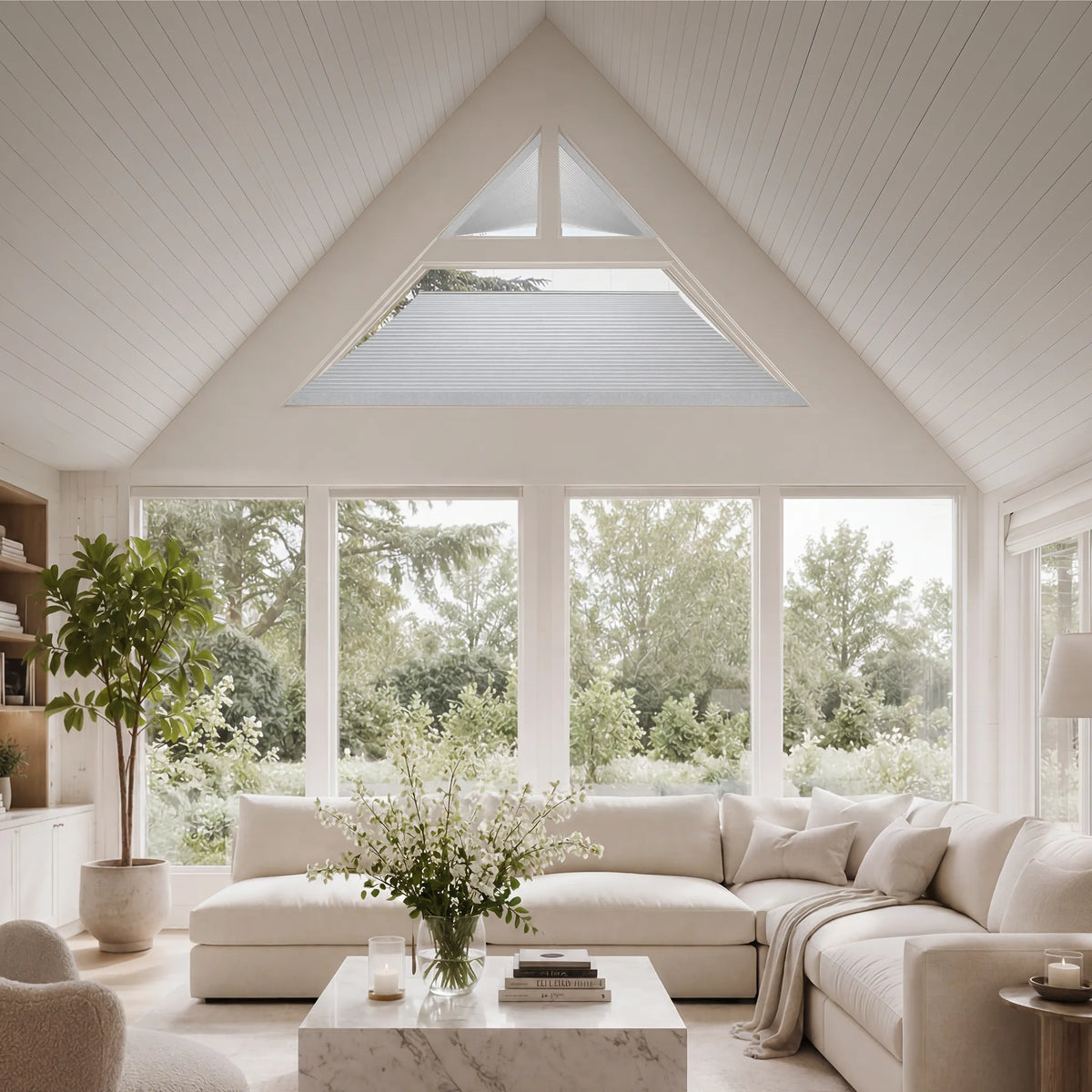 A bright, modern living room with a large white sectional sofa, a marble coffee table, and a tall potted plant, set against a white paneled wall and a Motorized Isosceles Trapezoid Cellular Shades looking out to green trees.
