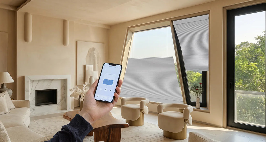 A person holds a smartphone showing a smart home app screen in a modern beige living room. The room features large black-framed windows with partially drawn gray Motorized Isosceles Trapezoid Cellular Shades, a fireplace, and neutral-toned furniture.
