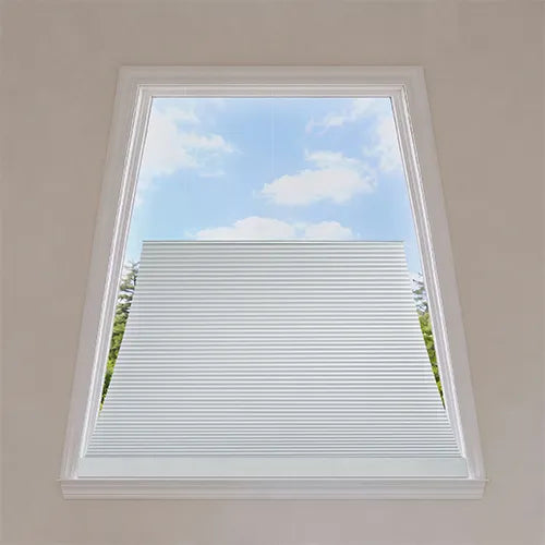 White Motorized Isosceles Trapezoid Cellular Shades installed on a tall, angled skylight window, with a blue sky and clouds visible outside.