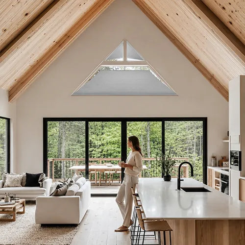 A woman in a white outfit stands in a bright, modern open-plan kitchen and living space with a large triangular skylight, wood-beamed ceiling, black-framed windows, and a white island. The room is surrounded by a forest view.