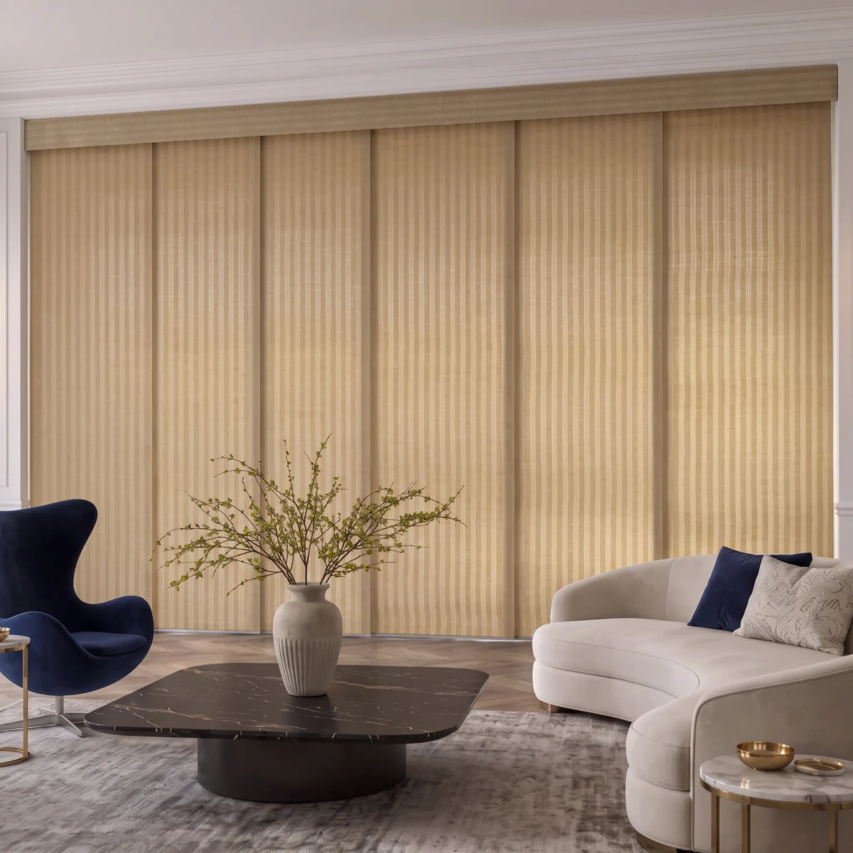 A staged living room interior featuring a large wall of beige Motorized Light Filtering Panel Track Blinds behind a curved cream sofa, dark marble coffee table, blue accent chair, and gray rug.