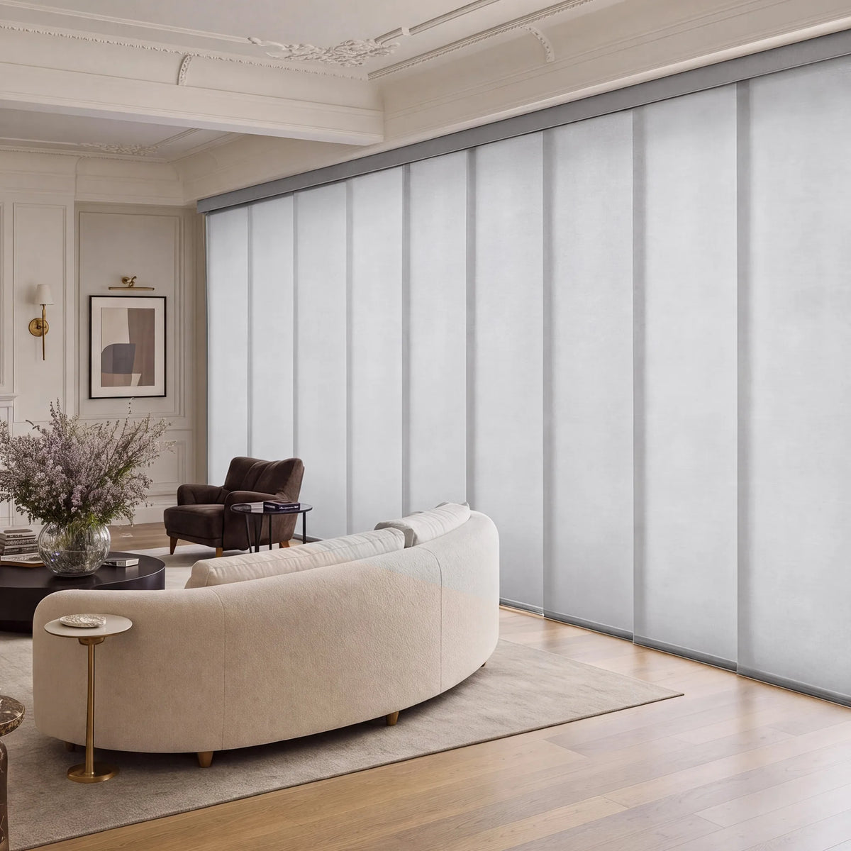 A modern living room with a curved cream sofa, dark brown armchair, and a large wall of frosted Motorized Light Filtering Panel Track Blinds in light gray. The room features light wood floors, a beige rug, and elegant white ceiling moldings.