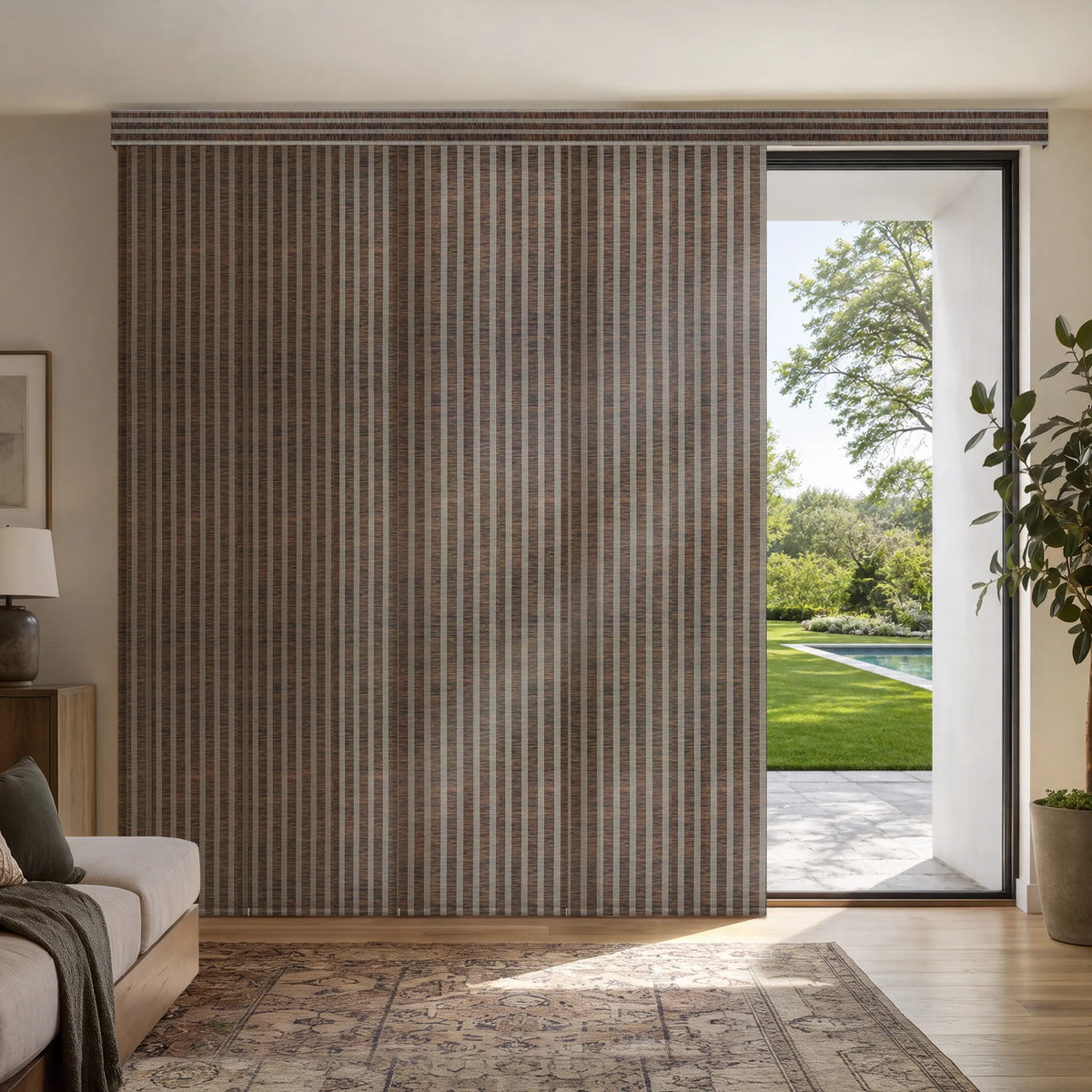 A large Motorized Light Filtering Panel Track Blinds in brown, beige, and gray tones, shown installed in a modern living room next to an open glass door revealing a green outdoor patio and pool.