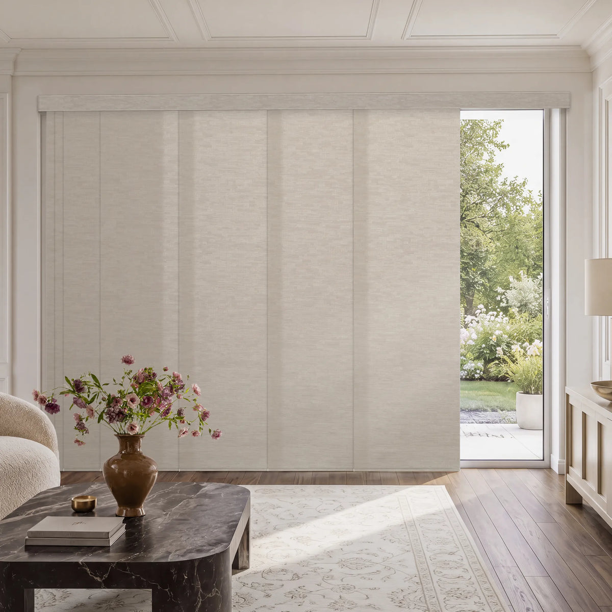 A set of tall, light beige textured Motorized Light Filtering Panel Track Blinds installed on a wall in a bright, modern living room with a marble coffee table and an open sliding door revealing a garden view.