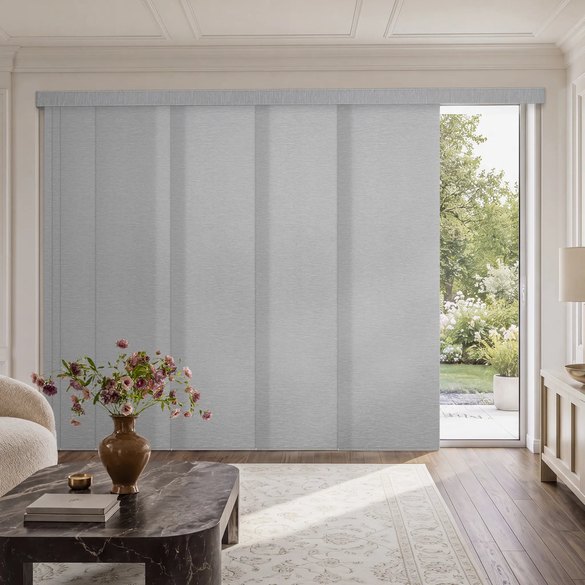 A light gray fabric Motorized Light Filtering Panel Track Blinds installed on a large window in a bright, modern living room with a marble coffee table and a view of a green garden outside.