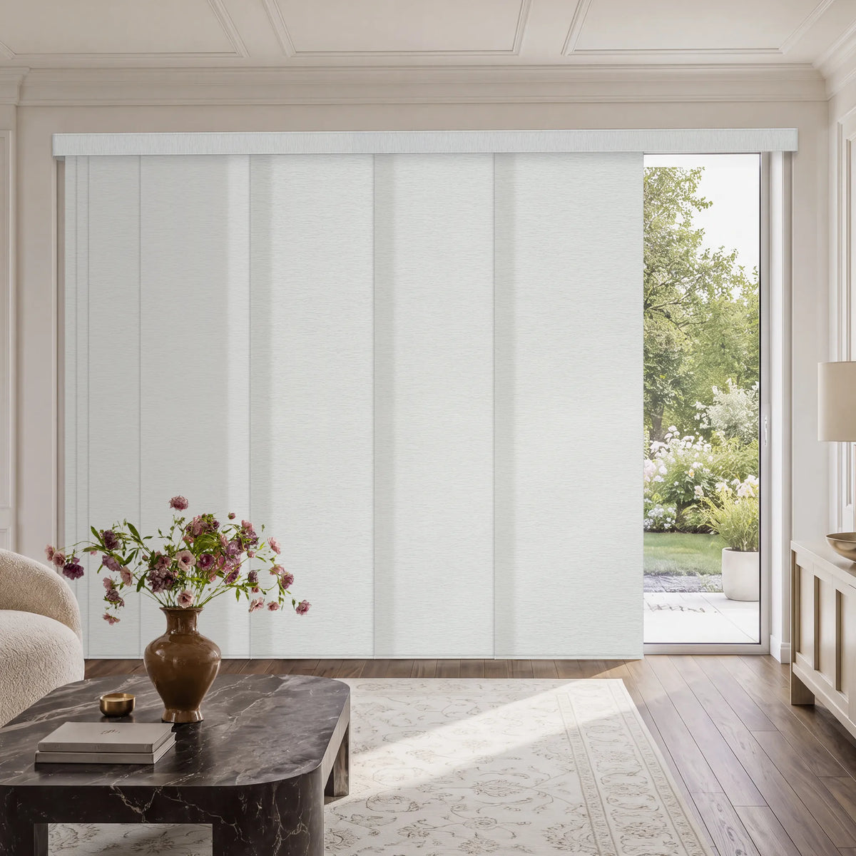 A set of white Motorized Light Filtering Panel Track Blinds covering a large sliding glass door in a bright, modern living room with a marble coffee table and a view of a green garden outside.
