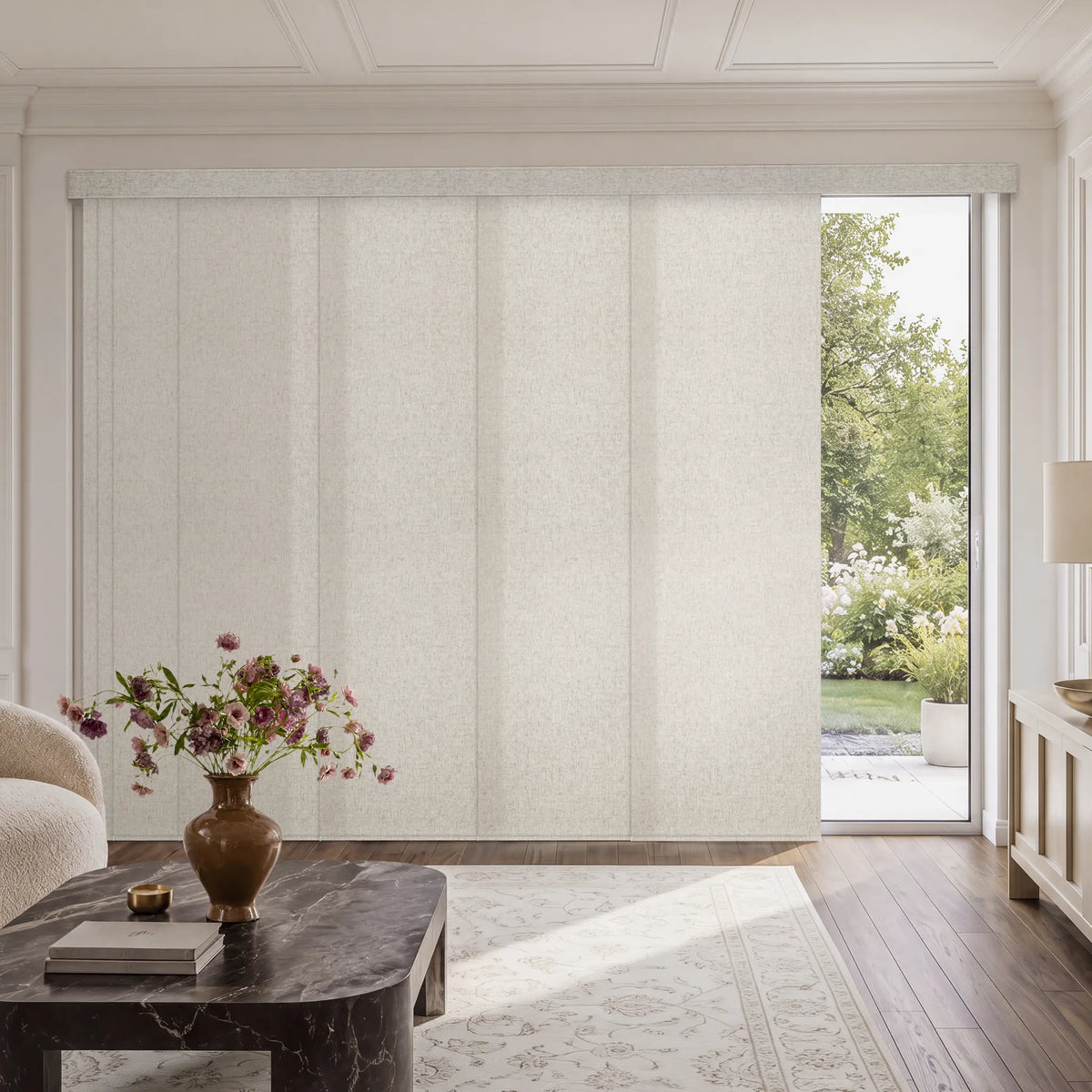 A set of tall, cream-colored textured Motorized Light Filtering Panel Track Blinds covering a large sliding glass door in a bright, modern living room with a marble coffee table and a view of a green garden outside.