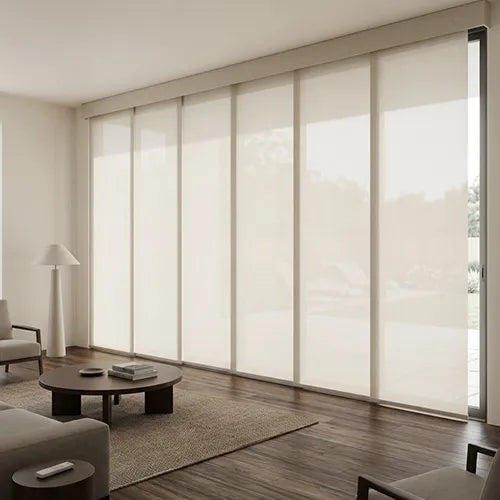 A modern living room with a large sliding set of beige Motorized Panel Track Blinds partially drawn, installed on a tall window wall. The room features dark wood floors, a low round coffee table, a white floor lamp, and neutral-toned furniture.