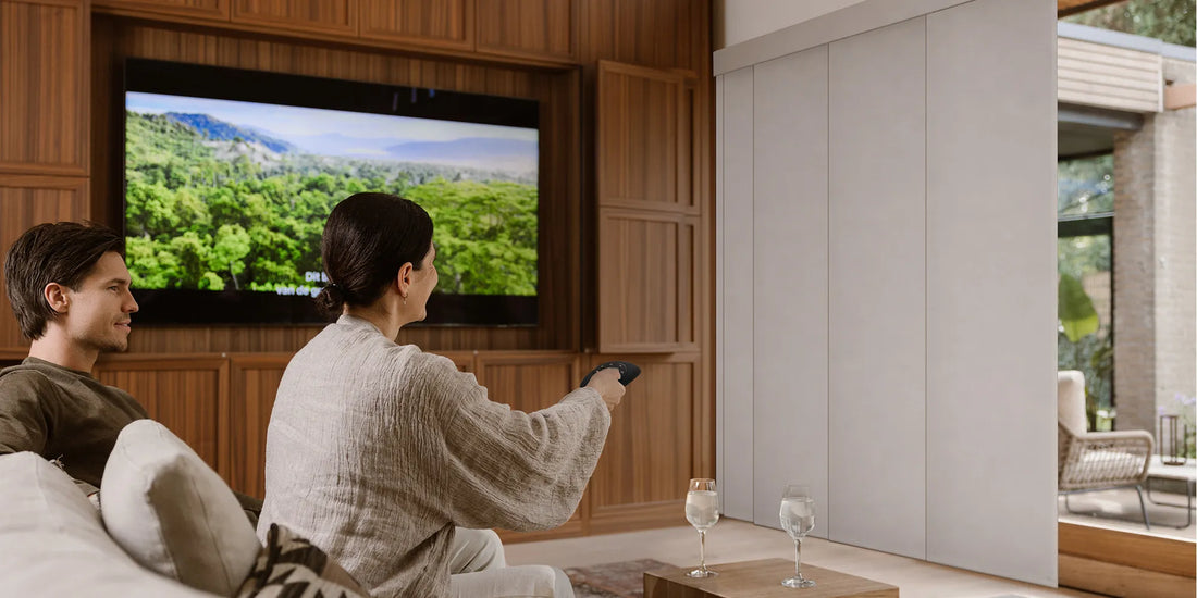 A man and a woman sit on a couch in a modern living room, watching a large wall-mounted television displaying a lush green landscape. The room features warm wood paneling, a white sliding door, and two wine glasses on a small table in the foreground.