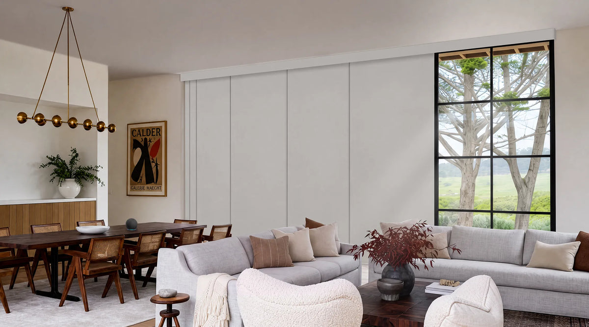 A modern living room interior featuring a light gray sectional sofa, a matching armchair, a dark wood coffee table with a vase of branches, and a dining area with a wooden dining table and chairs. A large black-framed window on the right shows trees outside, and a tall framed art print hangs on the beige wall.