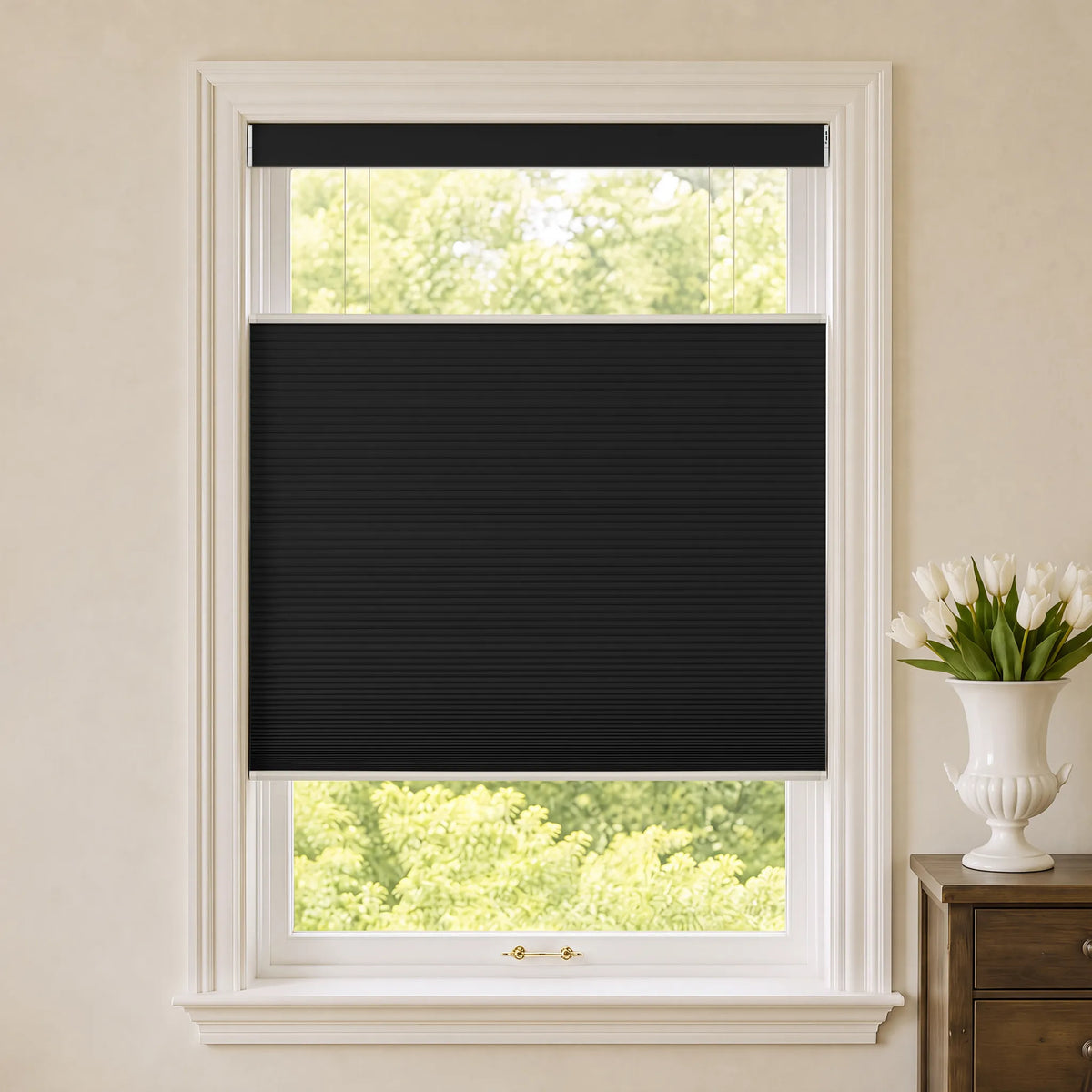 A black Motorized Top Down Bottom Up Blackout Cellular Shades  partially drawn over a white-framed window, with a cream wall, green foliage outside, and a white vase of tulips on a wooden side table to the right.