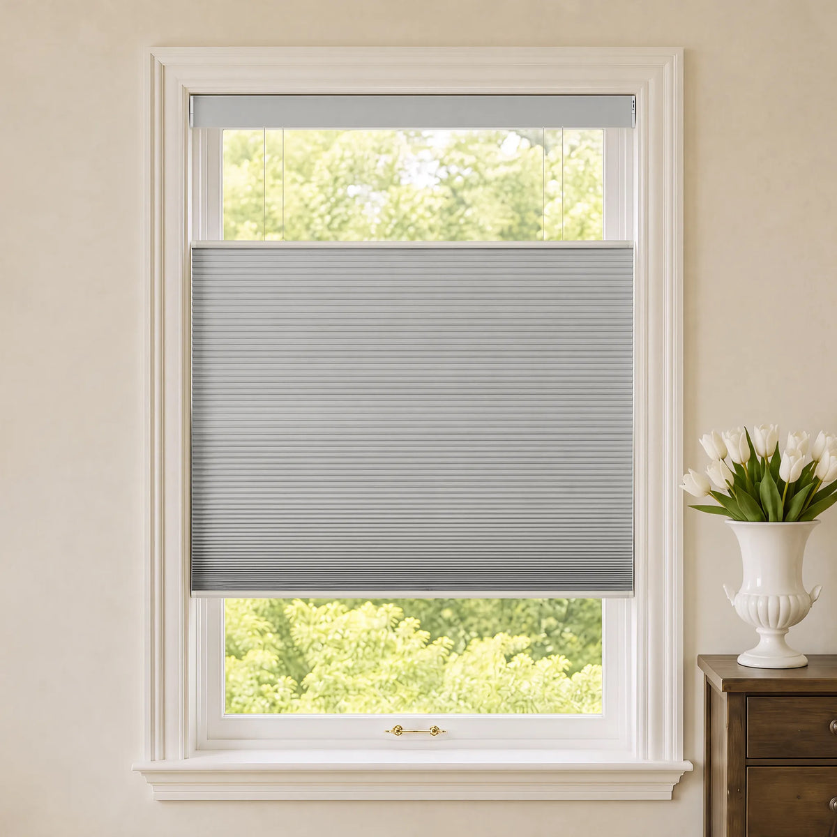 A gray Motorized Top Down Bottom Up Blackout Cellular Shades  partially drawn across a white-framed window, with a cream-colored wall, green trees outside, and a white vase of tulips on a wooden side table to the right.