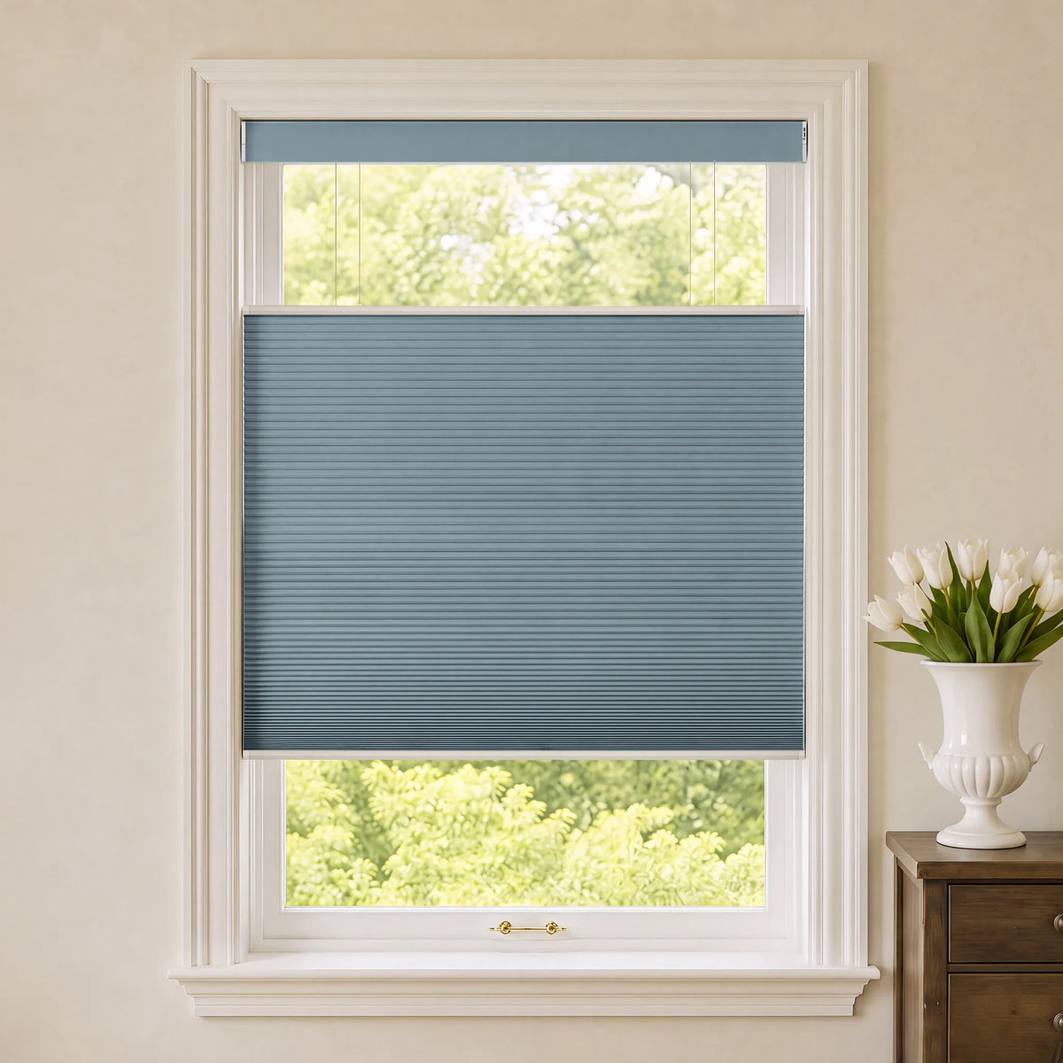 A blue-gray Motorized Top Down Bottom Up Blackout Cellular Shades  window blind shown installed in a white-framed window, with a view of green trees outside and a white vase of tulips on a wooden side table to the right.