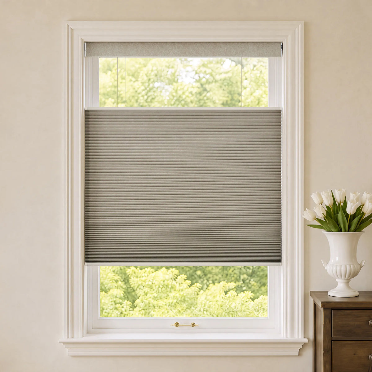 A coffee Motorized Top Down Bottom Up Blackout Cellular Shades partially drawn across a white-framed window, with a beige wall, green outdoor foliage visible, and a white vase of tulips on a wooden side table to the right.