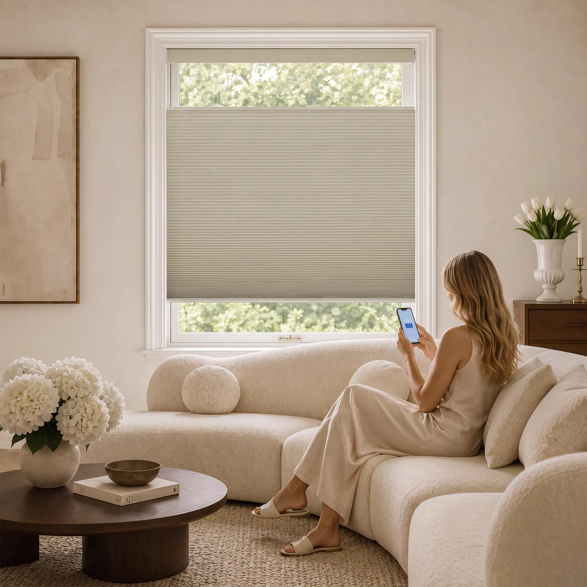 A woman sits on a cream sectional sofa in a bright living room, looking at her phone. A light beige Motorized Top Down Bottom Up Blackout Cellular Shades partially drawn over a white-framed window behind her.