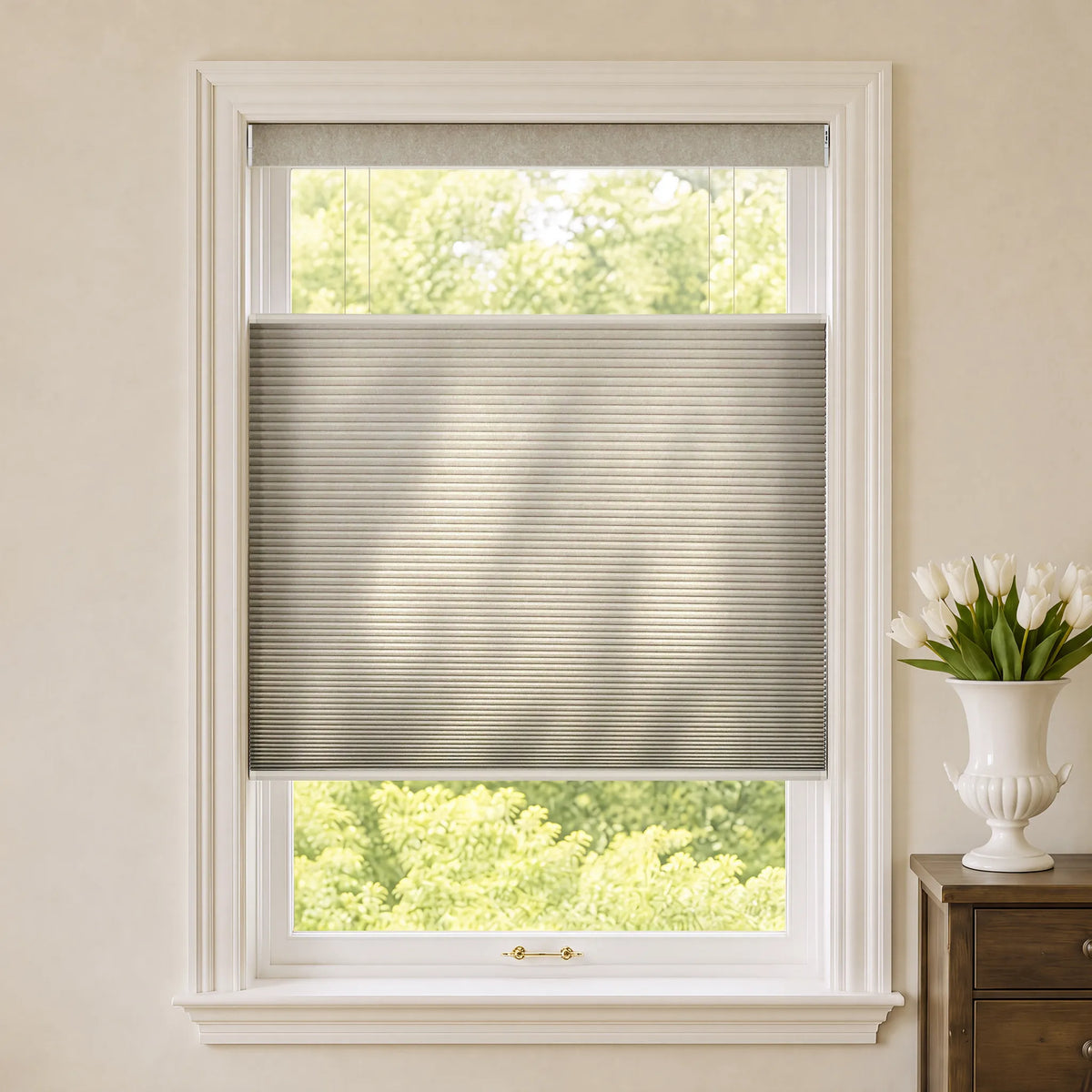 A gray Motorized Top Down Bottom Up Cellular Shades shown installed in a white window frame, with a view of green trees outside and a white vase of tulips on a wooden side table to the right.