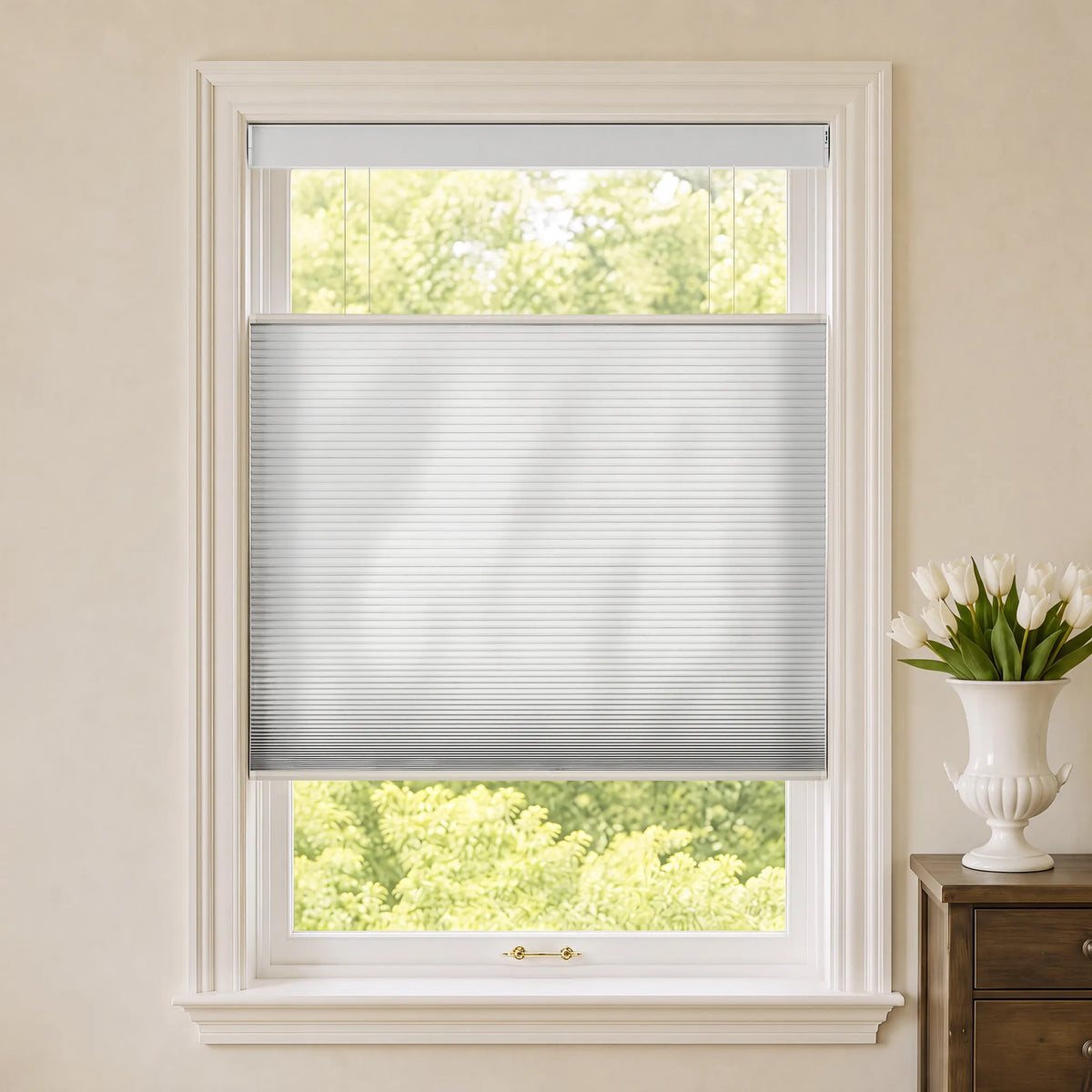 A white Motorized Top Down Bottom Up Cellular Shades partially drawn across a white-framed window, with a softly blurred green outdoor scene visible behind it. The room has beige walls, a white vase of tulips on a wooden side table to the right, and the window is centered in the frame.