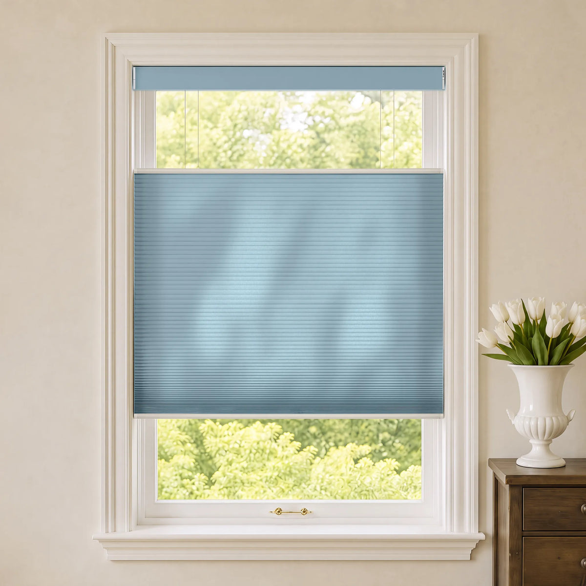 A blue Motorized Top Down Bottom Up Cellular Shades  partially drawn over a white-framed window, with a view of green trees outside and a white vase of tulips on a wooden side table to the right.