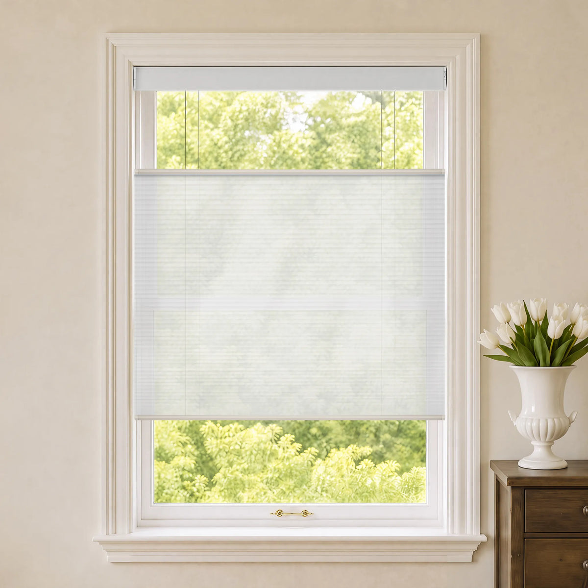 A sheer white Motorized Top Down Bottom Up Cellular Shades  partially drawn over a white-framed window, with a view of green trees outside and a white vase of tulips on a wooden side table to the right.