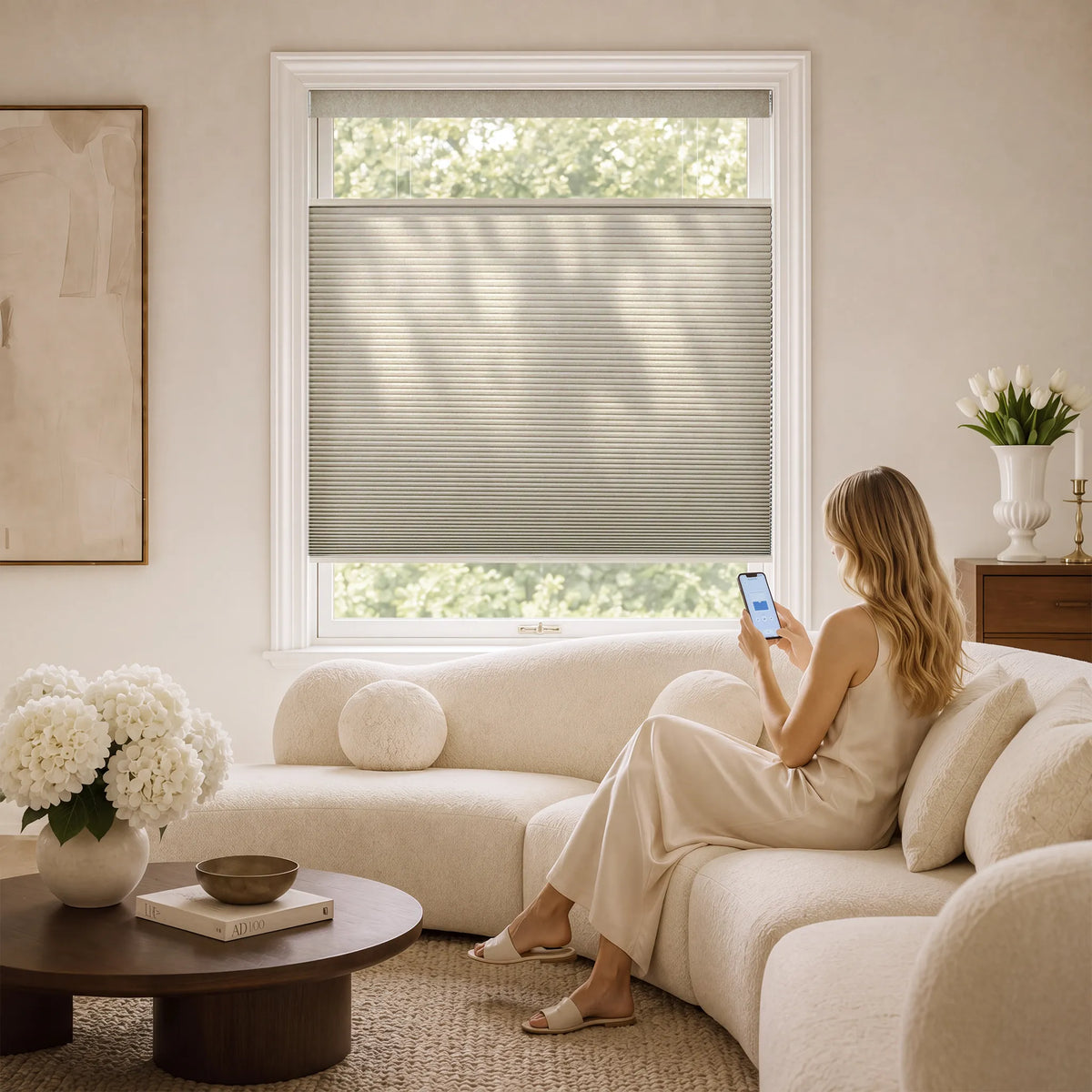 A woman sits on a cream sectional sofa in a bright living room, using her phone beside a window with a gray Motorized Top Down Bottom Up Cellular Shades drawn down. The room has warm neutral walls, a round wooden coffee table with white flowers, and a vase of tulips on a sideboard.