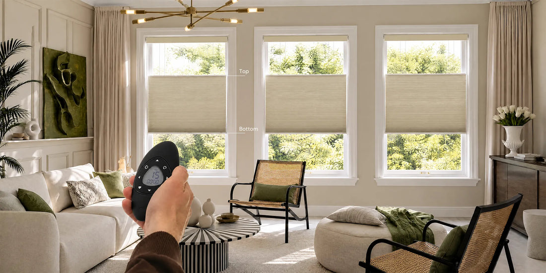 A hand holds a black remote control in a bright, modern living room with three windows featuring beige Motorized Top Down Bottom Up Cellular Shades . The room is styled with a white sofa, green accent pillows, a rattan armchair, and neutral decor.