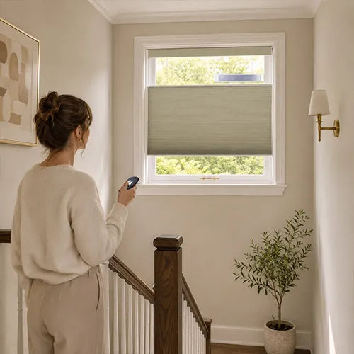 A woman stands in a hallway near a window with a Motorized Top Down Bottom Up Cellular Shades  partially drawn, holding a remote control. The room has beige walls, a wooden staircase railing, a wall sconce, and a potted plant.
