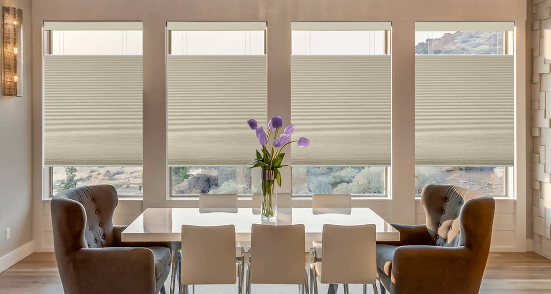A dining room with four large windows fitted with beige Motorized Top Down Bottom Up Cellular Shades , a white dining table, four upholstered chairs, and a vase of purple flowers in the center.