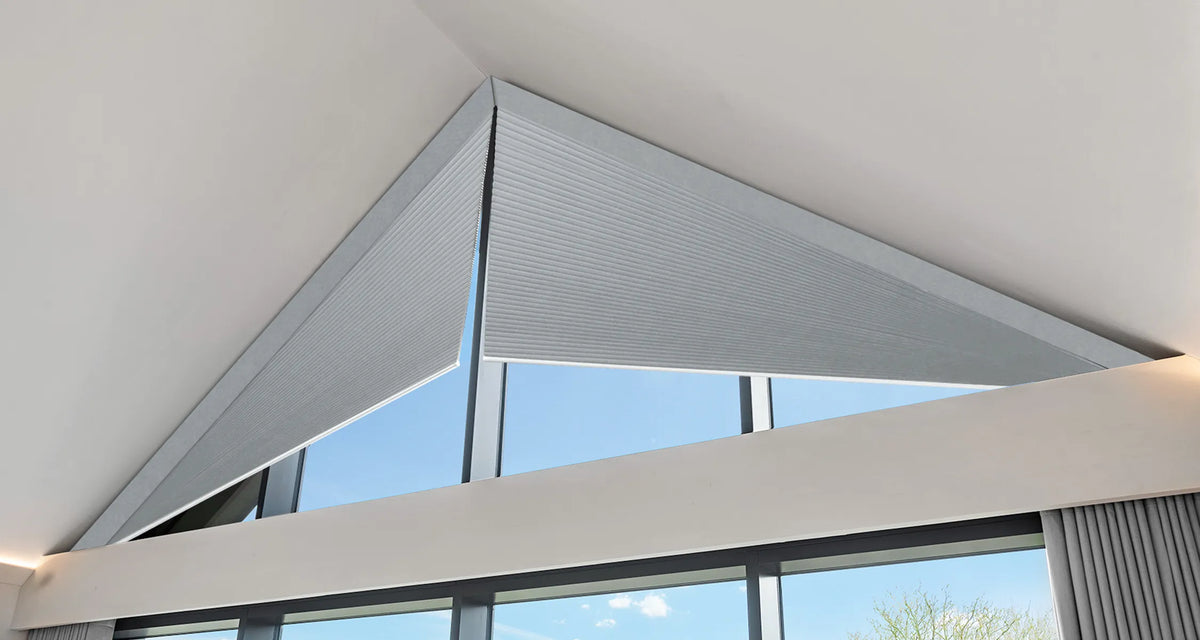 A triangular gray roller blind or shade mounted on a large angled window in a modern interior with white walls and a view of blue sky.