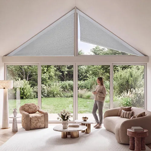 A woman in a white outfit stands in a bright, modern open-plan kitchen and living space with a large triangular skylight, wood-beamed ceiling, black-framed windows, and a white island. The room is surrounded by a forest view.