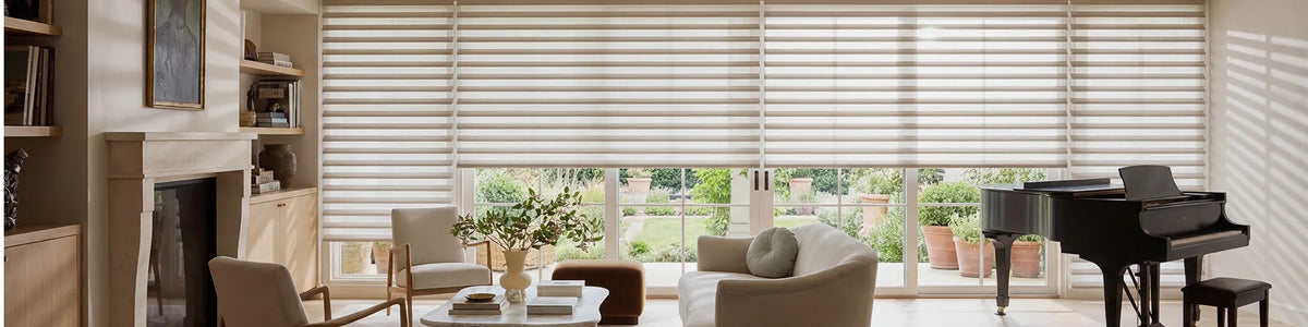A large interior room with beige roller blinds covering tall windows, with a black grand piano, upholstered chairs, a fireplace, and a garden view outside.